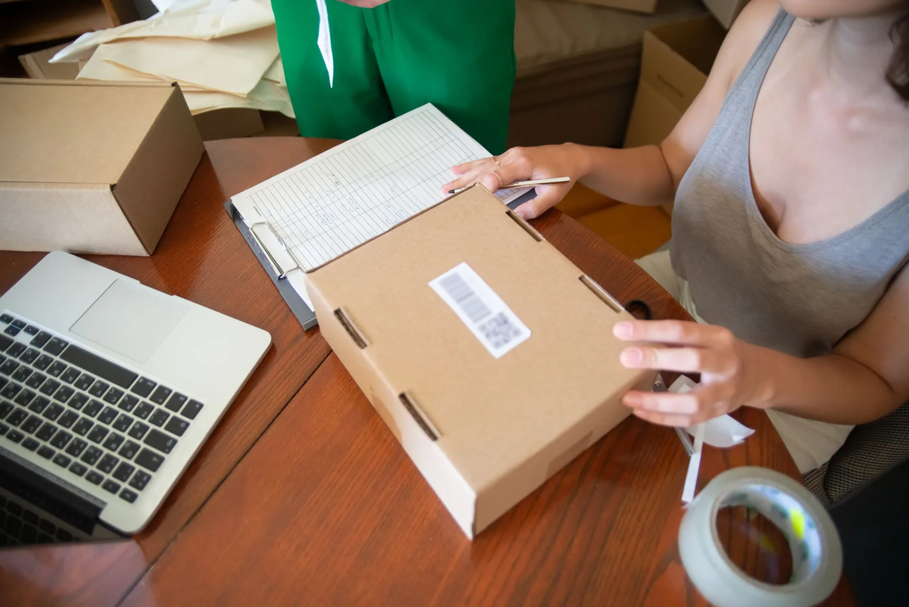 An ecommerce operator preparing a cardboard order beside a laptop and packing list.
